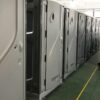 Row of gray portable toilet stalls in a facility hallway, doors closed and lined up along a green floor with a yellow safety line.