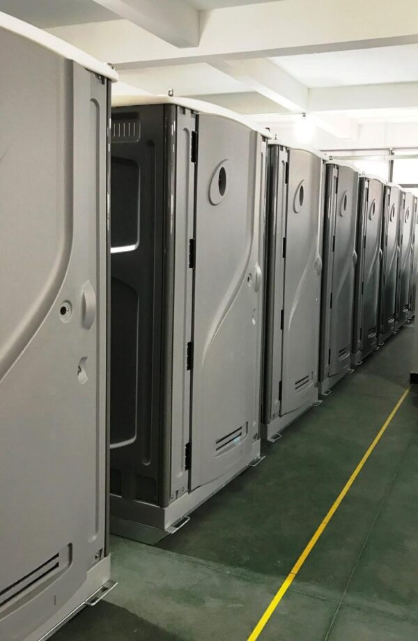 Row of gray portable toilet stalls in a facility hallway, doors closed and lined up along a green floor with a yellow safety line.
